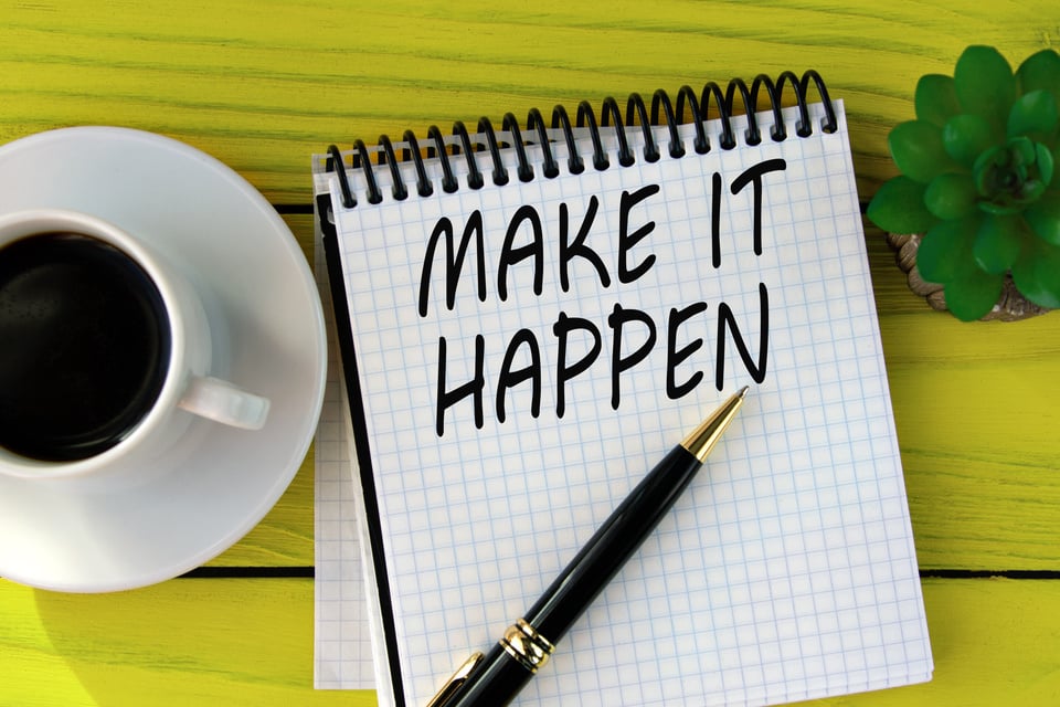 MAKE IT HAPPEN - words on a white sheet on a yellow wooden background with a pen and a cup of coffee